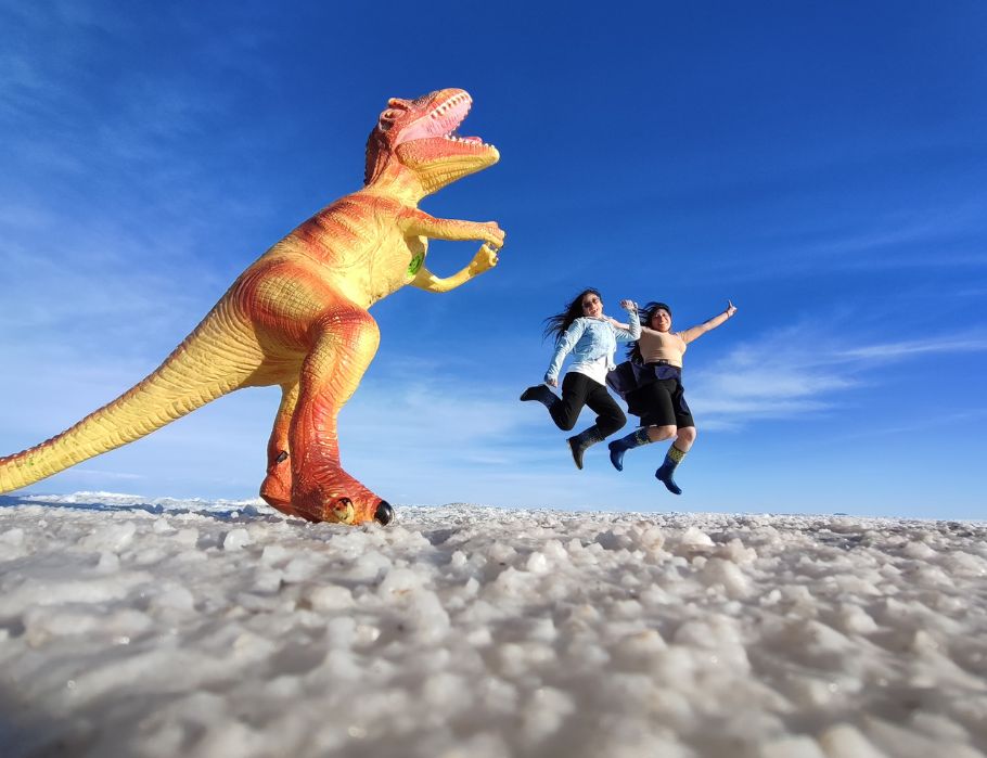 Dos viajeras saltando en el Salar de uyuni mientras un dinosaurio las persigue
